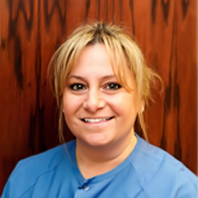 Registered Dental Hygienist Tara Smiling In Scrubs In Front Of Wood Wall